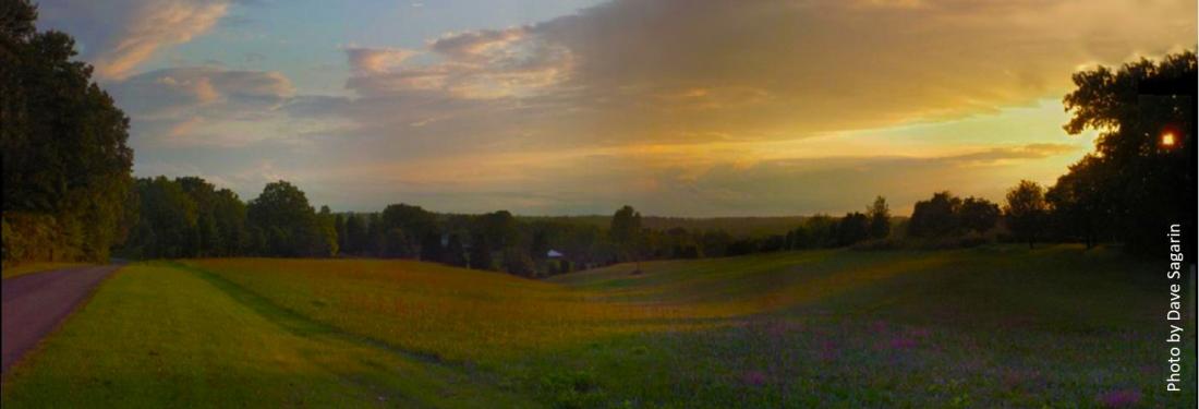 a large field in the foreground with clouds and sun setting in distance