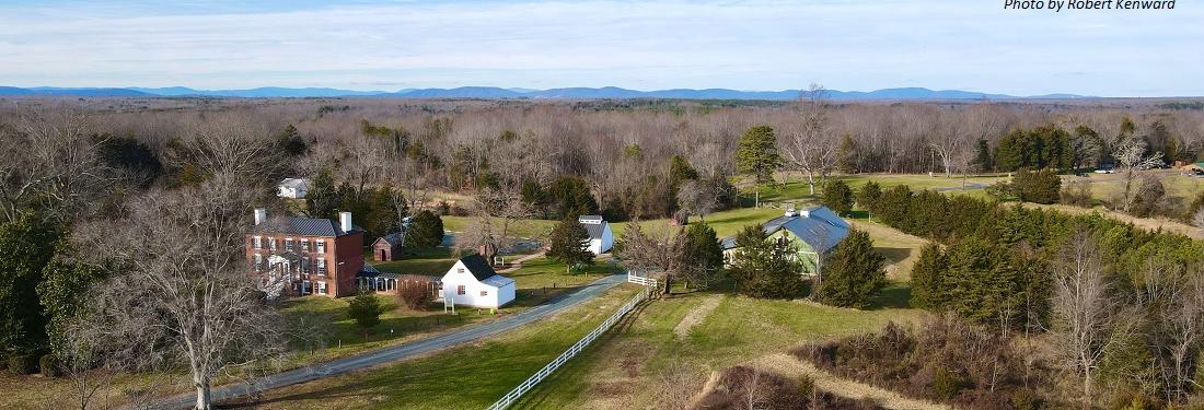 a house and barn are seen from the air. mountains and clouds are in the distance.