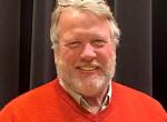 a smiling man with a beard wears a red sweater 
