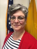 a woman with short hair wearing a striped shirt and red cardigan stands in front of 2 flags